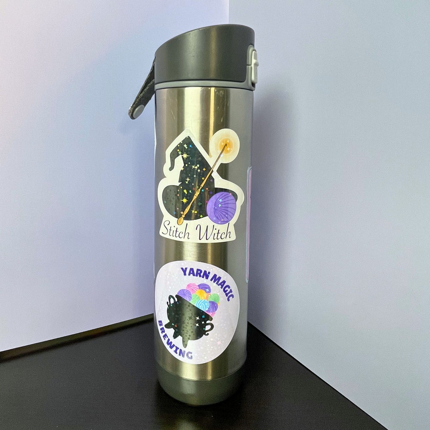 Metallic water bottle with 'Stitch Witch' and 'Yarn Magic Brewing Co.' labels on a black surface.